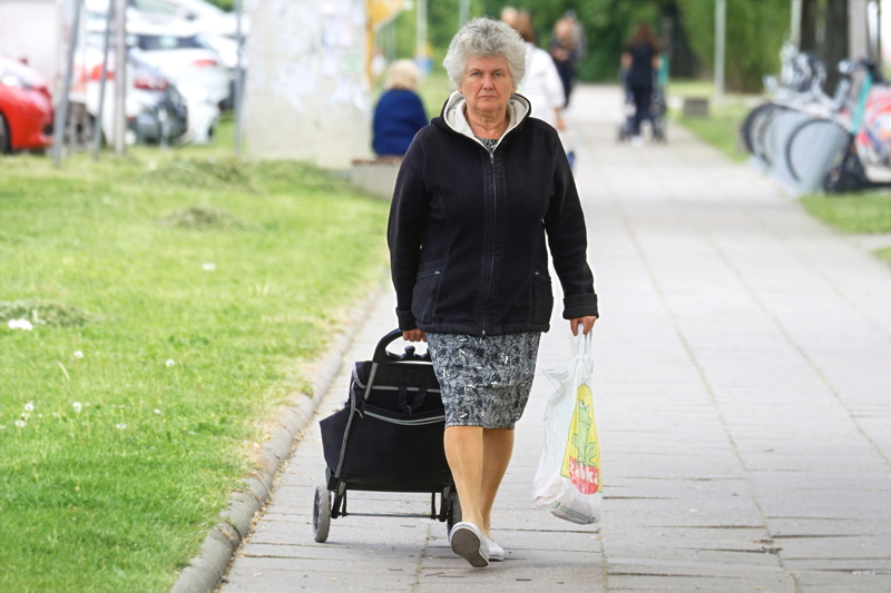 11 - An elderly woman walking down a sidewalk while pulling a black rolling shopping cart and carrying a white plastic bag